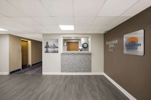 Un'area della reception di un hotel con un orologio, un quadro incorniciato che raffigura un panorama urbano e un cartello con la scritta 'Rodeway Inn' e 'Suburban Studios by Choice Hotels'.
