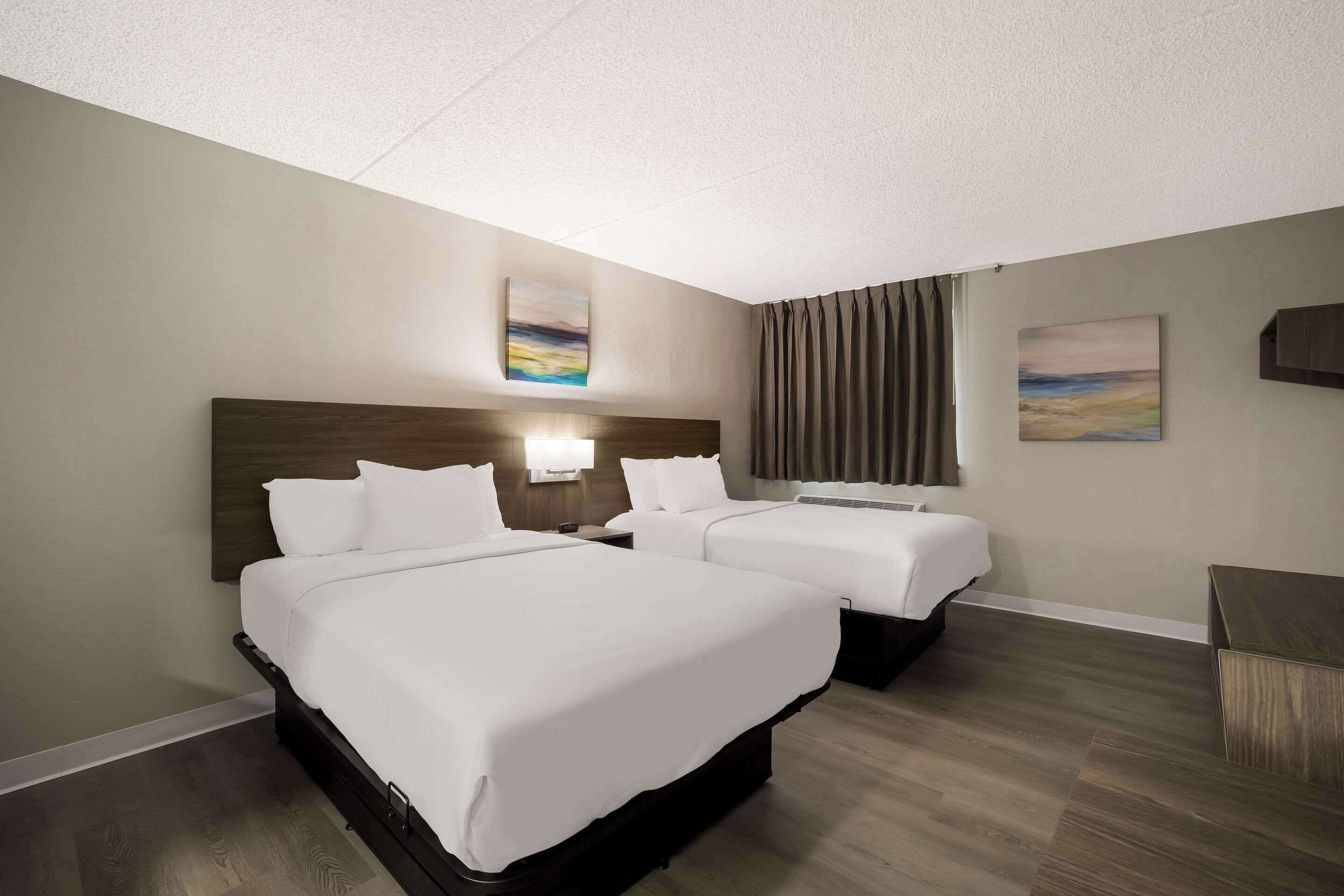 A hotel room with two beds, a headboard, a nightstand, a lamp, and two paintings on the wall.