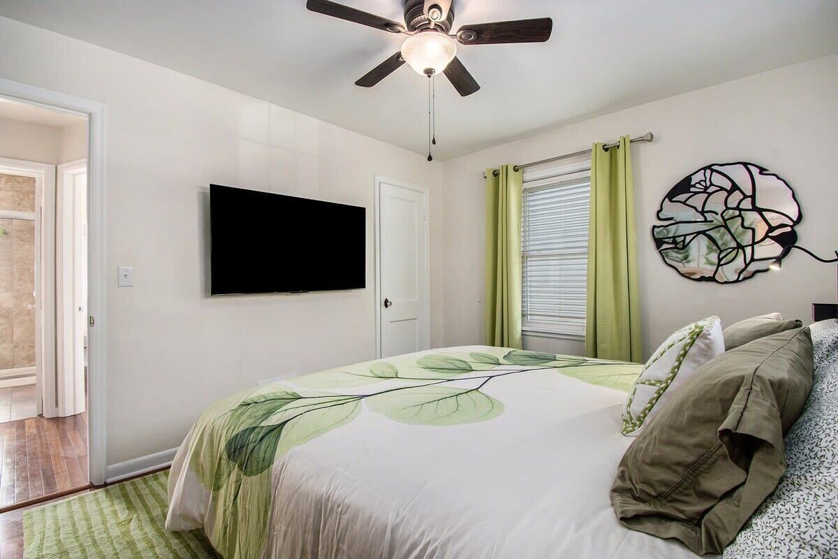 A bedroom with a bed, a ceiling fan, a flat-screen TV, a window with curtains, and a wall art piece.