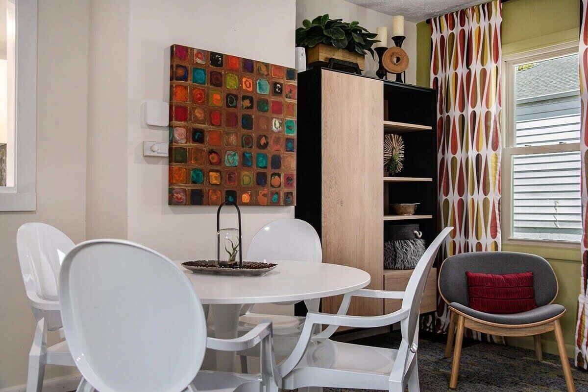 A dining area with a round table and white chairs, a colorful mosaic wall, a wooden cabinet with shelves, and a window with patterned curtains.