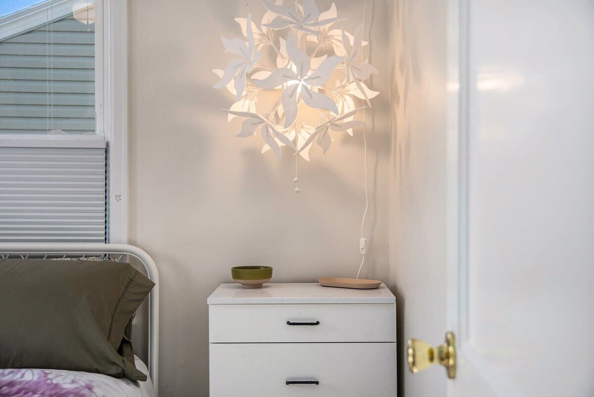 Een slaapkamer met een witte nachtkastje, een groene kom en een witte lamp met bladvormige lampenkappen.