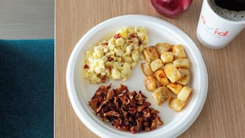 Un plato con huevos revueltos, papas asadas y tocino, una manzana roja y una taza de café sobre una mesa de madera.