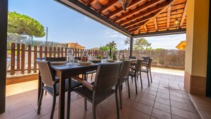 Een terras buiten met een houten plafond, betegelde vloer en rieten stoelen.
