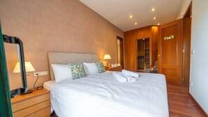 Een net opgemaakt bed met witte lakens, een houten nachtkastje met een lamp, en een houten garderobe met een spiegel.