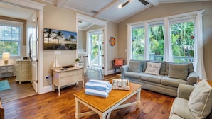 A spacious living room with a wooden floor, a ceiling fan, a sofa, and a coffee table with towels. - Pool - Beach Bellini by SeaBreeze Vacation (Holmes Beach)