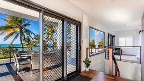 A modern hotel room with a balcony overlooking the ocean, featuring a wooden floor, a bed with a view, and a small potted plant. - Beautifully Renovated Large Home on Lammermoor Beach. (Yeppoon)