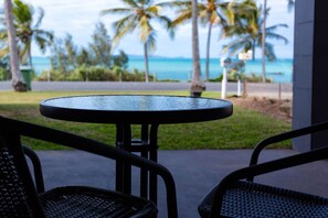 Une table ronde avec un cadre en métal, deux chaises en osier et une vue sur une route bordée de palmiers, avec un plan d’eau en arrière-plan.