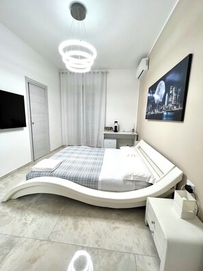 A modern bedroom with a curved bed, a mounted TV, a painting of a cityscape, and a ceiling light fixture. - Circeo Boutique Rooms  (San Felice Circeo)