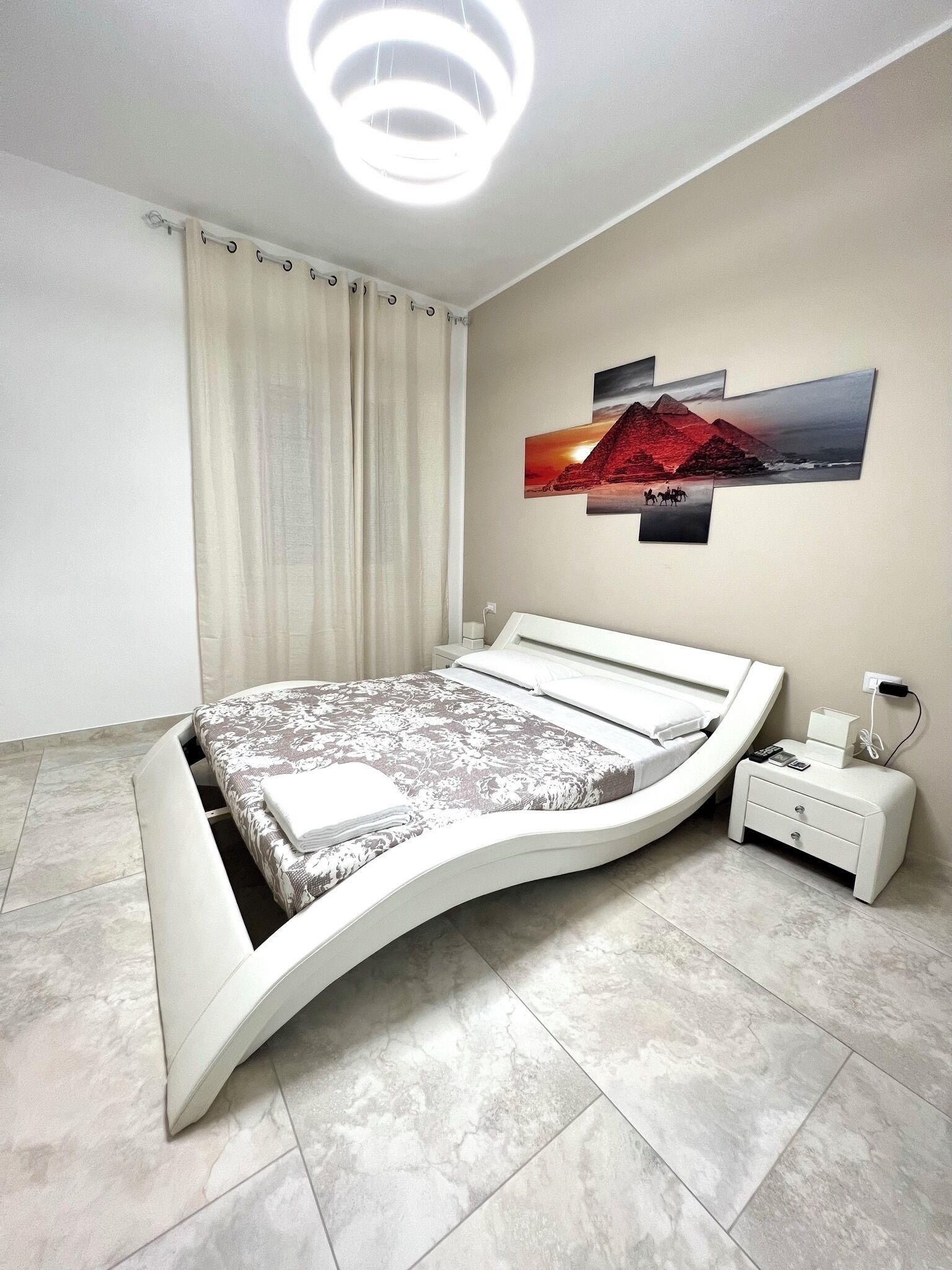 A modern bedroom with a curved bed, a nightstand, a wall-mounted light fixture, and a framed picture of a sunset over a pyramid.