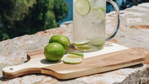 A clear glass pitcher with lime slices on a stone ledge, accompanied by whole and sliced limes on a wooden board, overlooking a coastal landscape with trees and the sea.