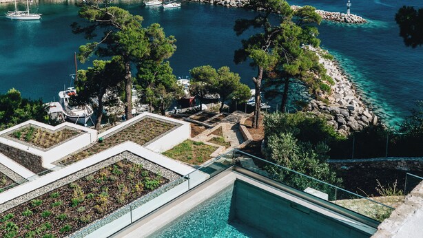 A pool with clear water, surrounded by a stone deck and greenery, overlooking a coastal area with boats and rocky terrain.