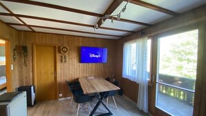 A room with a wooden interior, a flat-screen TV mounted on the wall, a dining table with chairs, and large windows with curtains. - Cozy 2BR Apartment with Mountainview* Free Parking (Flums)