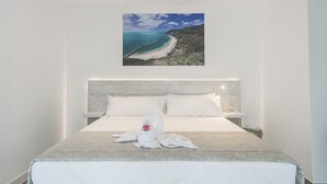 Una cama bien tendida con una manta blanca y un almohadón decorativo con forma de cisne, un cabecero y un cuadro enmarcado de un paisaje costero encima.