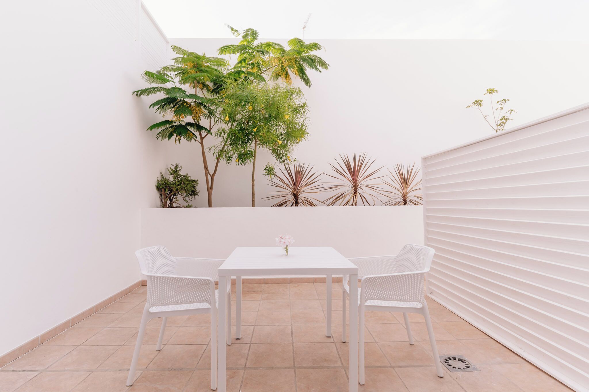 Eine kleine Terrasse mit einem weißen Tisch und zwei Stühlen, umgeben von Pflanzen und einer weißen Mauer.
