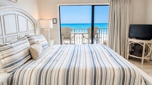 A hotel room with a bed, a TV, a balcony with chairs, and a view of the ocean.