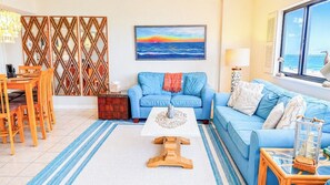 A living room with a blue sofa, a dining table with chairs, a coffee table, and a view of the ocean through a window. - Shalimar 2B (North Myrtle Beach)