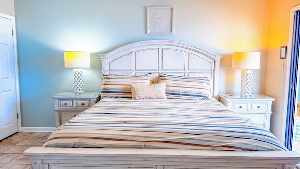 A bed with a striped bedspread, two bedside tables with lamps, and a wall-mounted picture. - Shalimar 2B (North Myrtle Beach)