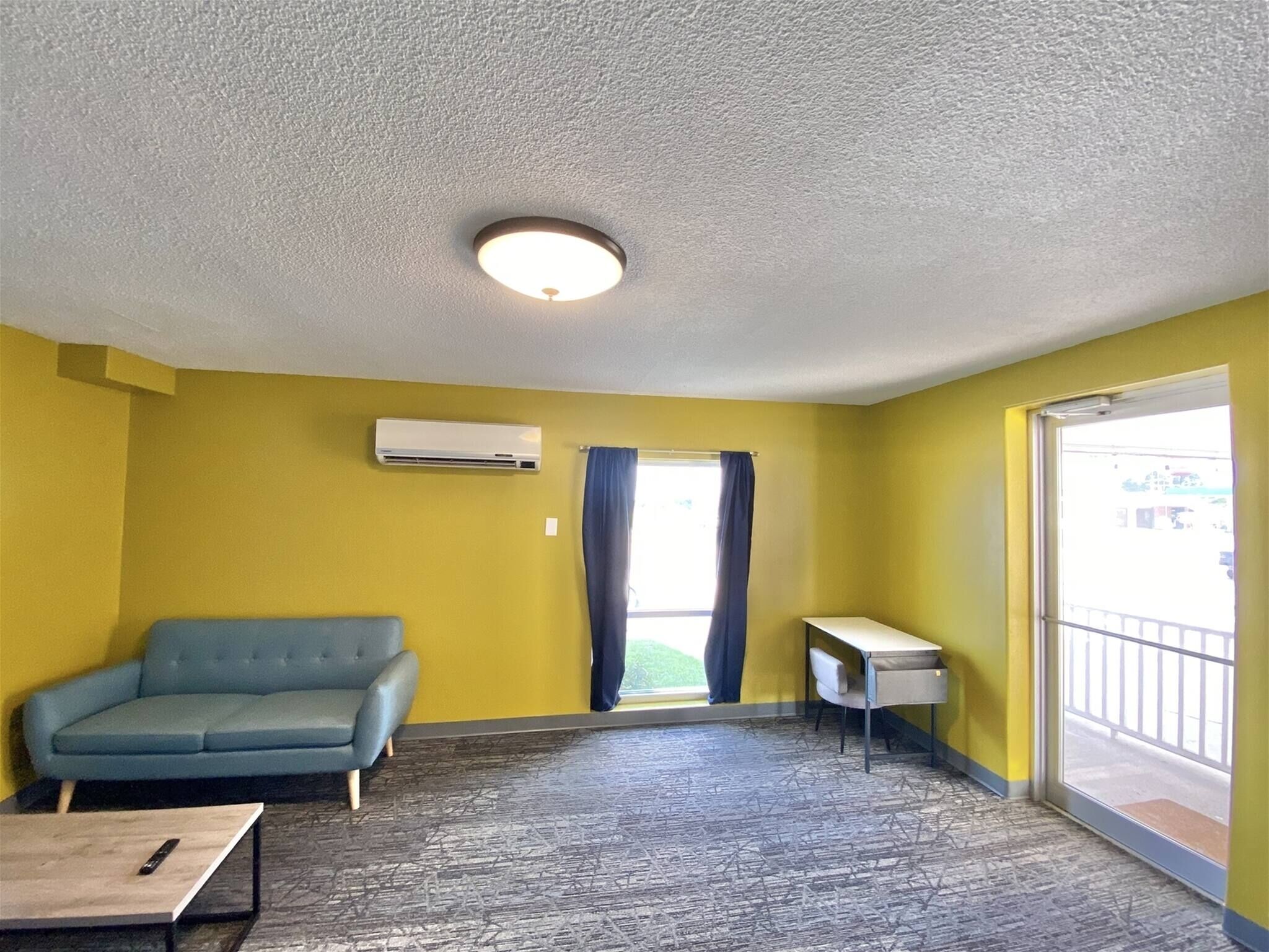 A room with a blue sofa, a small table, an air conditioning unit, and a window with curtains.