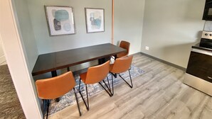 Uma sala de jantar com uma mesa de madeira escura, quatro cadeiras laranjas, dois quadros abstratos e uma cozinha com fogão.