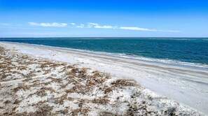 A beach with white sand and sparse vegetation, leading to a calm sea under a clear sky.