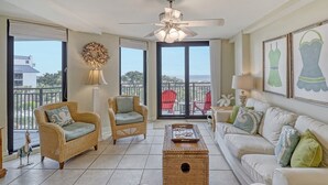 Amplia sala de estar con muebles de mimbre, ventilador de techo y ventanas grandes con vista al océano.