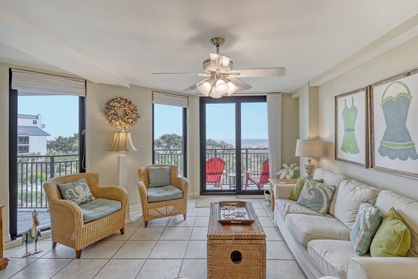 A spacious living room with wicker furniture, a ceiling fan, and large windows offering a view of the ocean.