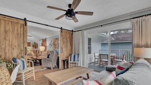 A spacious living room with a wooden floor, a ceiling fan, a large sofa, a coffee table, and a sliding glass door leading to an outdoor patio. - Little Cottage by the Sea | Hot Tub & Walk to Beach (Isle of Palms)