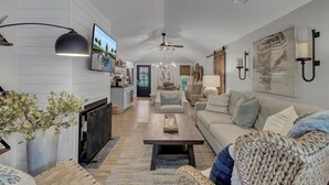 A modern living room with a sofa, coffee table, and a TV. There is a kitchen area in the background with cabinets and a countertop. - Little Cottage by the Sea | Hot Tub & Walk to Beach (Isle of Palms)