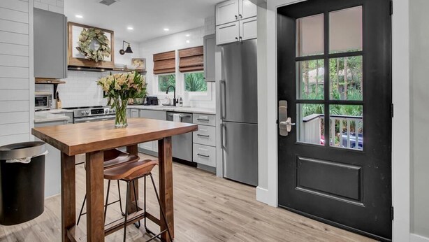 A modern kitchen with a wooden table, bar stools, a refrigerator, and a black door with windows. - Little Cottage by the Sea | Hot Tub & Walk to Beach (Isle of Palms)