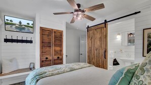 A bedroom with a bed, a ceiling fan, wooden wardrobe, a mounted TV, and a bathroom with a sink and mirror. - Little Cottage by the Sea | Hot Tub & Walk to Beach (Isle of Palms)