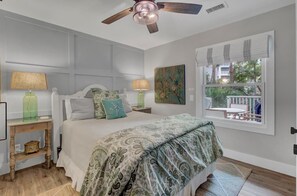 Une chambre à coucher avec un lit, deux tables de chevet, un ventilateur de plafond, une fenêtre donnant sur un beau paysage et un tableau accroché au mur.
