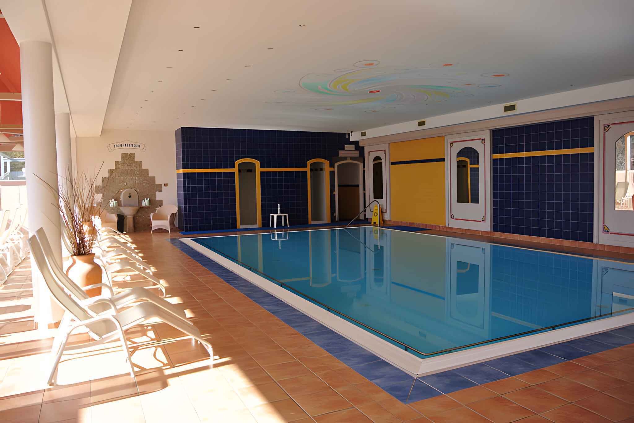 Una piscina con revestimiento de azulejos, tumbonas y una pared decorativa con una fuente.