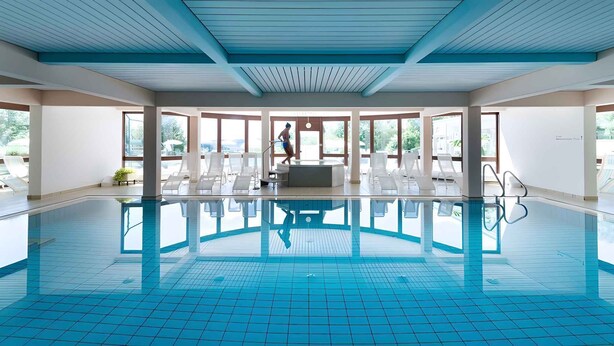 A large indoor swimming pool with clear blue tiles, a central hot tub, and sunlit seating areas.
