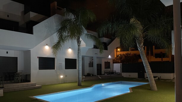 Zona de piscina exterior moderna amb una piscina ben il·luminada, envoltada per gespa artificial i palmeres.