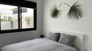Un dormitori amb un llit, una tauleta de nit, una finestra i dues fulles de palmera penjades a la paret.