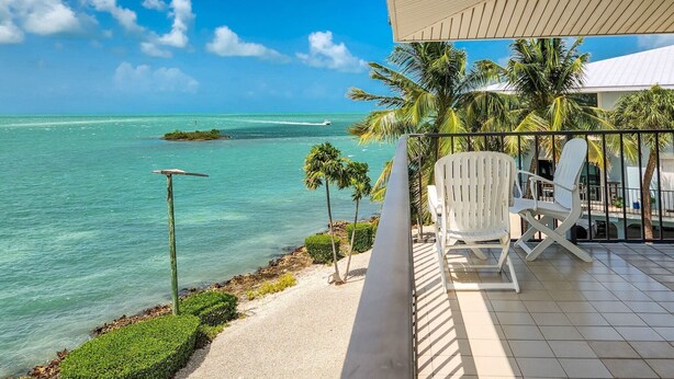 A balcony with white chairs, a view of the ocean, and palm trees.