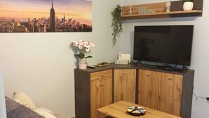 A living room with a sofa, a coffee table, a TV, a cabinet, a wall-mounted air conditioner, a cityscape painting, a welcome sign, and a potted plant.