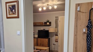 A modern living room with a sofa, a television, and a wooden cabinet. There is a door leading to another room with a lamp and a ceiling light fixture.