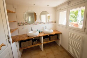 Een badkamer met twee wasbakken, houten planken, rieten manden, een betegelde spatwand en een raam met uitzicht.