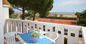 Een balkon met een tafel en stoelen, met uitzicht op een tennisbaan en strand.