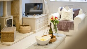 Uma sala de estar acolhedora com lareira, televisão, sofá com almofadas, um vaso com flores e uma travessa de madeira com frutas.