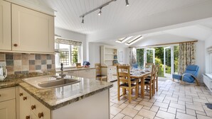 Una cocina amplia con una isla grande, una mesa de comedor de madera y sillas, un área de comedor con vista al jardín, y un salón con una mecedora azul.