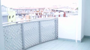 Een balkon met een geperforeerd metalen hekwerk, een betegelde vloer en een uitzicht op gebouwen in de verte.