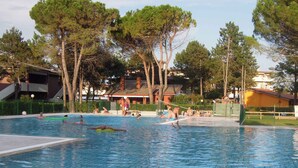 Ein Schwimmbecken mit Schwimmern und spielenden Personen, umgeben von Bäumen und Gebäuden.