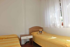 A single room with two beds, one with a striped bedspread and the other with a plain yellow bedspread. There is a small wooden nightstand with a drawer and a teddy bear on it. A window with white curtains is visible, allowing natural light into the room.