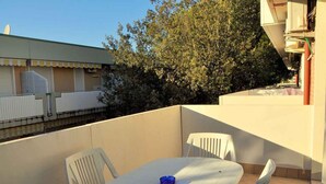 A balcony with a table and chairs, a tree, and a building in the background. - Holiday apartment mit Klimaanlage und Balkon (Bibione)