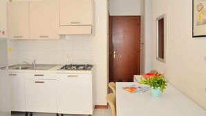 A compact kitchen with white cabinets, a gas stove, and a sink. There is a wooden door leading to another room, and a table with a plant and a magazine. - Holiday apartment mit Klimaanlage und Balkon (Bibione)