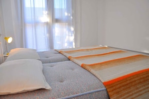 A hotel room with two beds, a nightstand with a lamp, and a window with sheer curtains.