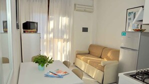 A compact living space with a kitchenette, a sofa, a dining table with a plant, and a TV on a cabinet. - Holiday apartment mit Klimaanlage und Balkon (Bibione)