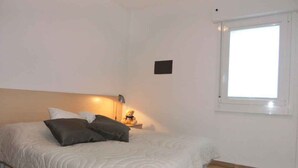 A bedroom with a bed, pillows, a bedside lamp, a small table, and a window. - Holiday apartment near the sea (Bibione)